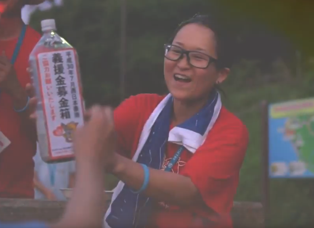 義援金募金箱とかかれた紙を貼り付けた2Lのペットボトルを持って、募金を笑顔で受け付けている中村さんの写真。